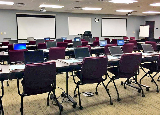 Photo of the training room with laptops on three rows of desks facing a podium and projector screens