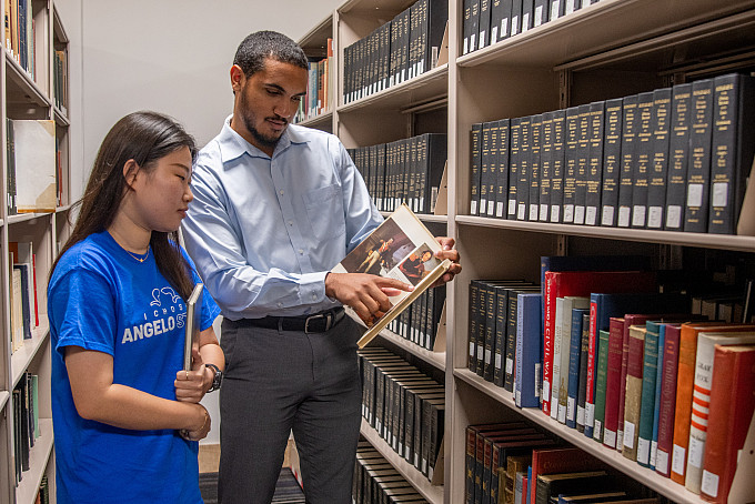 Students standing next to a bookshelf looking at a book together.