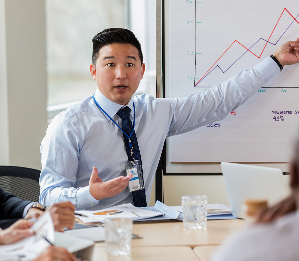 Hospital and Health Care Facilities Administration 1135118025 A serious businessman sits at a conference table with coworkers. He points to a graph behind him and gestures as he speaks.