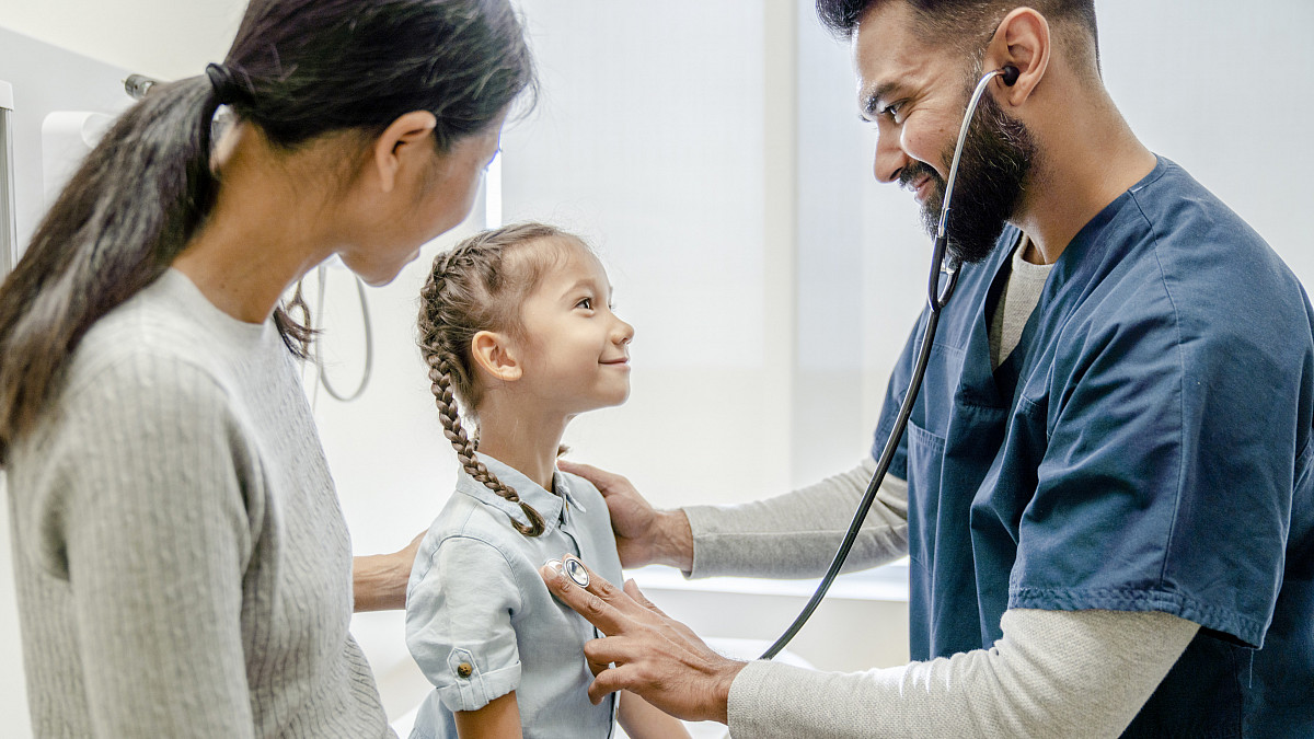 A male family nurse practitioner using a stethoscope to examine a child patient's chest.