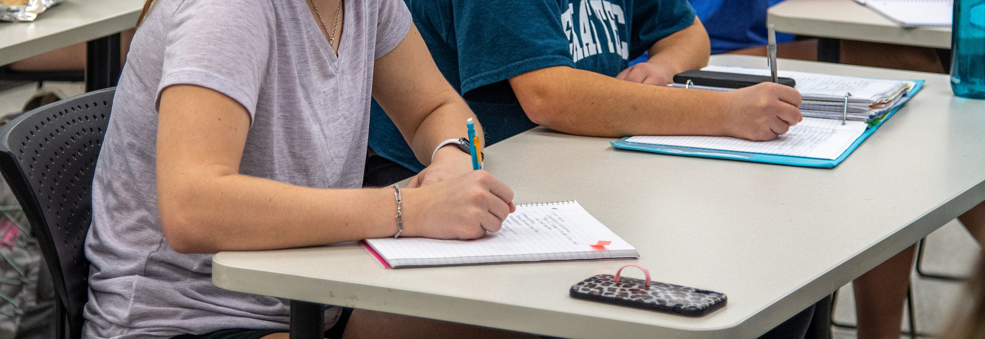 Students listen to the instructor and take notes with other students in the background.
