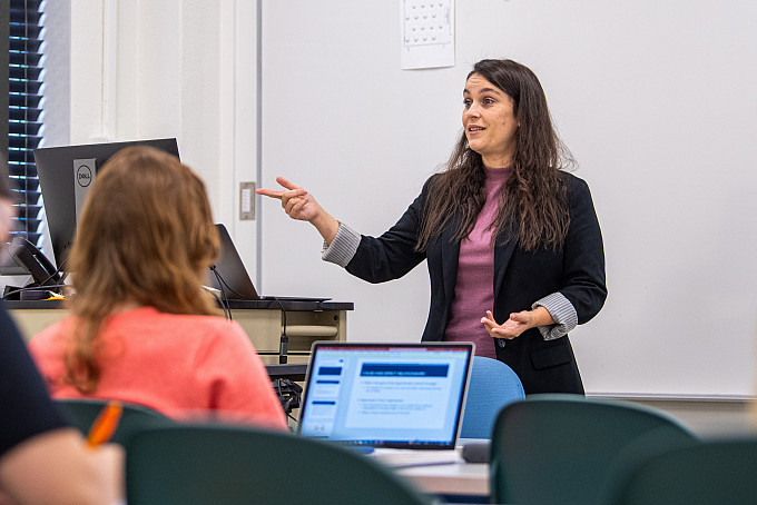 Professor speaks and points to the screen while students in the foreground listen and take notes.