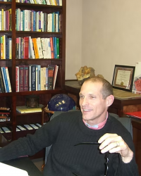 man sitting at a desk in front of a bookshelf, smiling and holding a pair of glasses